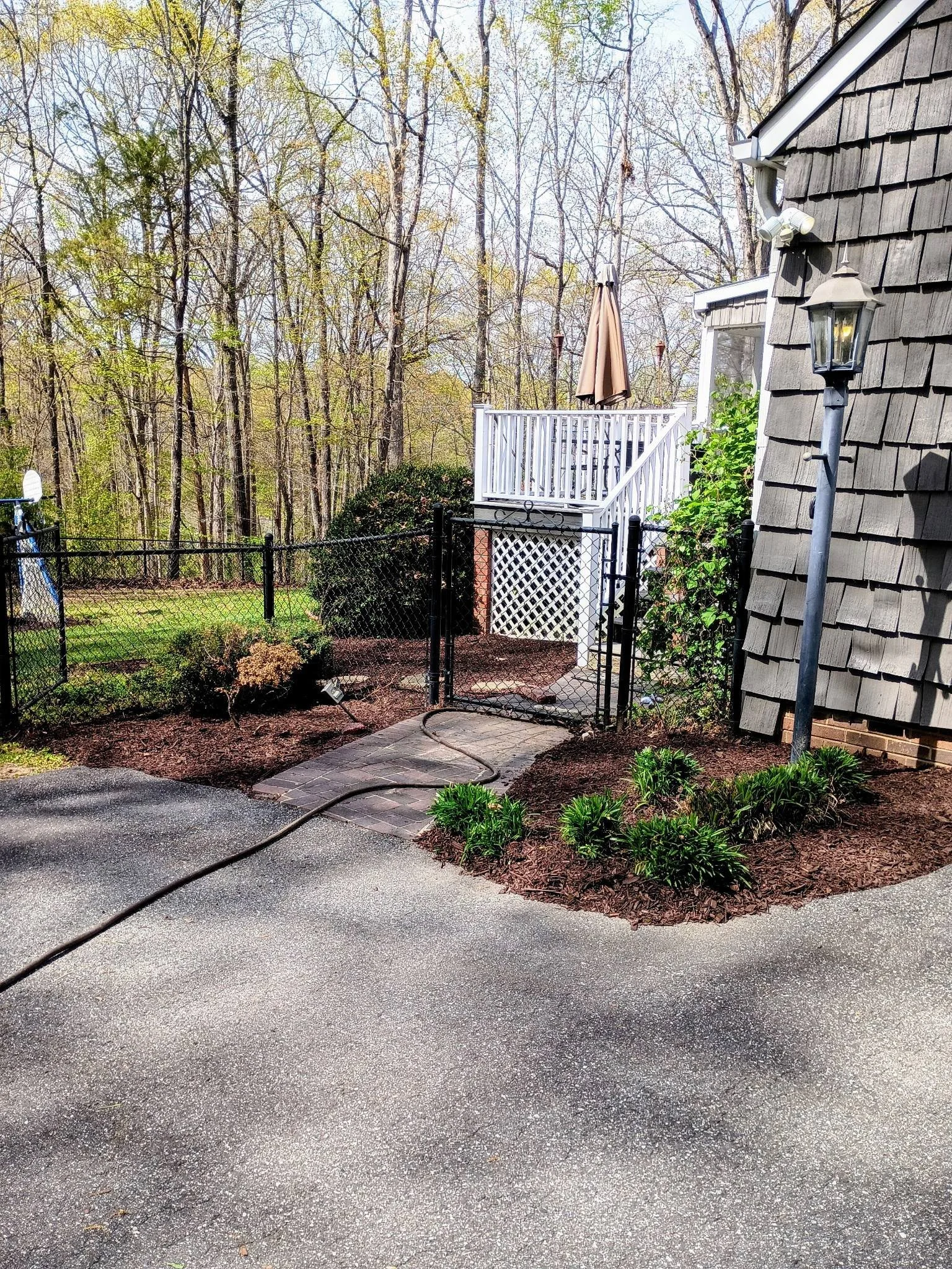 Backyard patio area with a black gated entrance, stone pathway, small garden beds with green plants and mulch, a white deck with a tan umbrella, a lamppost, and a wooded area in the background. After mulch