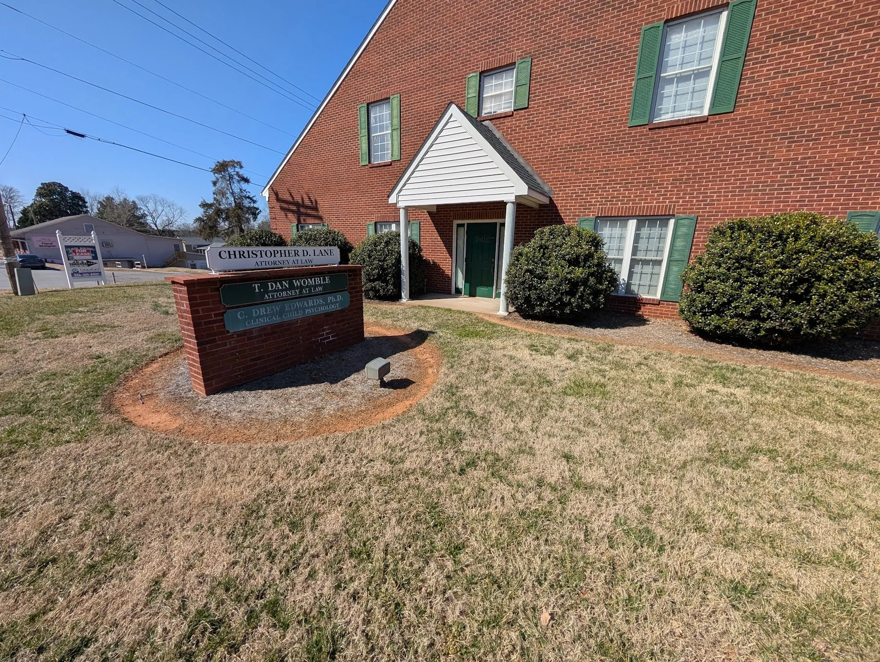 Red brick building with green shutters and a small porch with white columns. There is a sign in front for multiple attorneys, including Christopher D. Lane, T. Dan Womble, and C. Drew Edwards, located on a small grassy area.