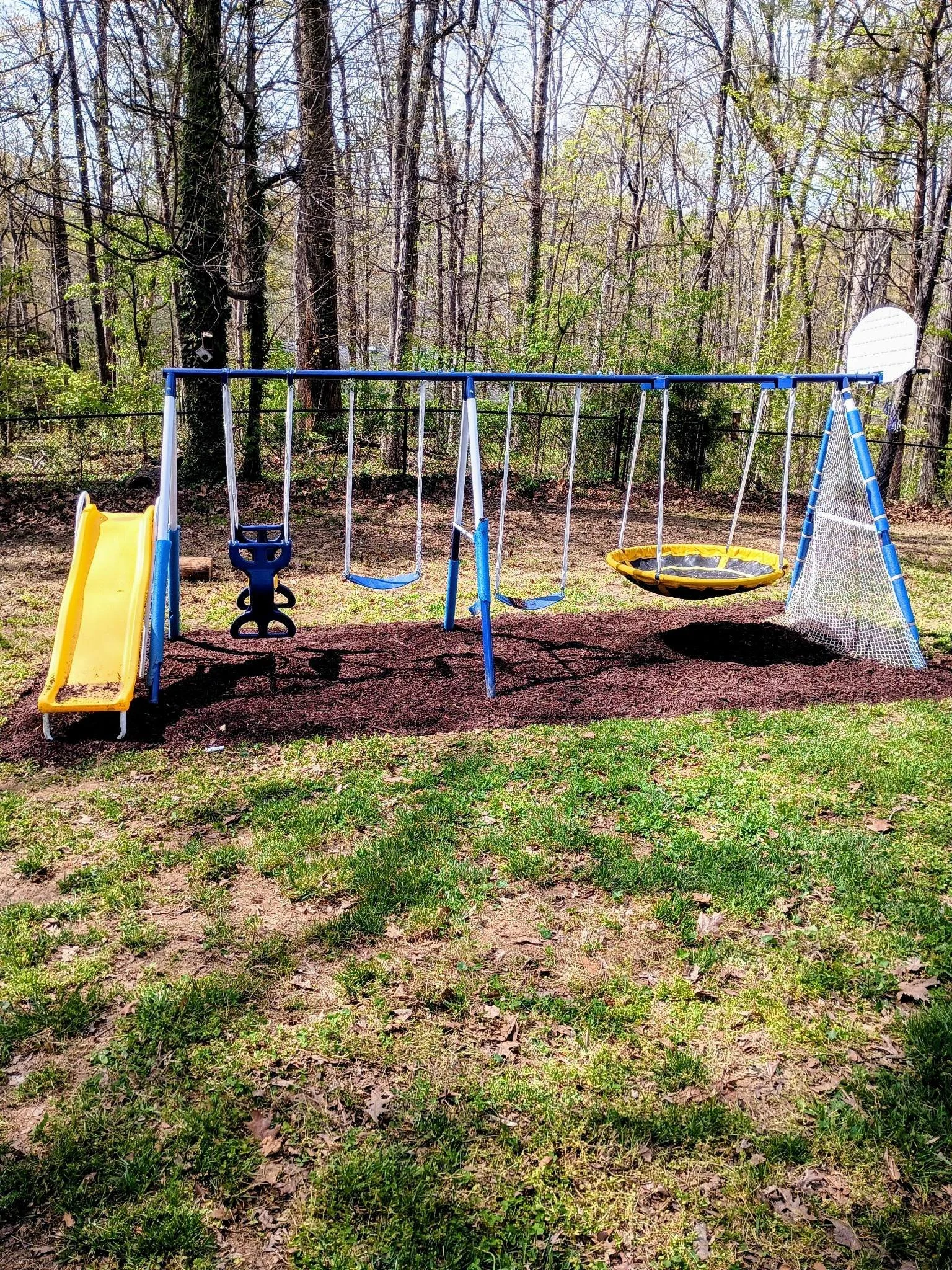 A children's outdoor playground set featuring a yellow slide, two blue swings, a yellow round swing, and a white netted side panel, set on a grassy yard with trees in the background. after mulch and string-trimming