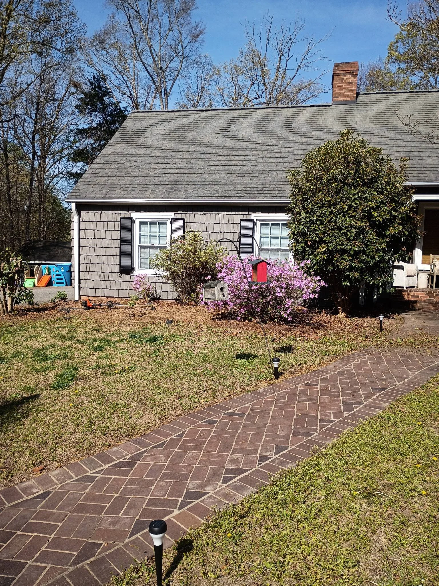 Front view of a house with a brick pathway, flowering bushes, a birdhouse, and outdoor toys on a sunny day. Before fresh mulch