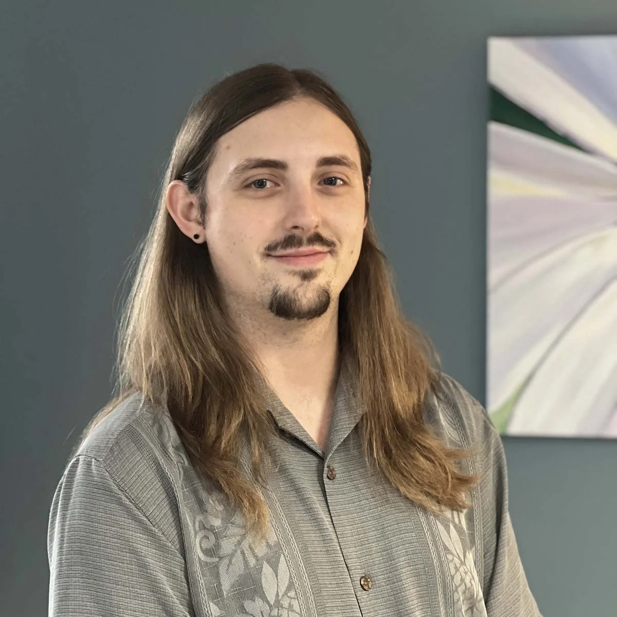 Portrait of a young man with long brown hair, a small mustache, and a goatee, wearing a gray patterned shirt, standing in front of a gray wall with abstract artwork.