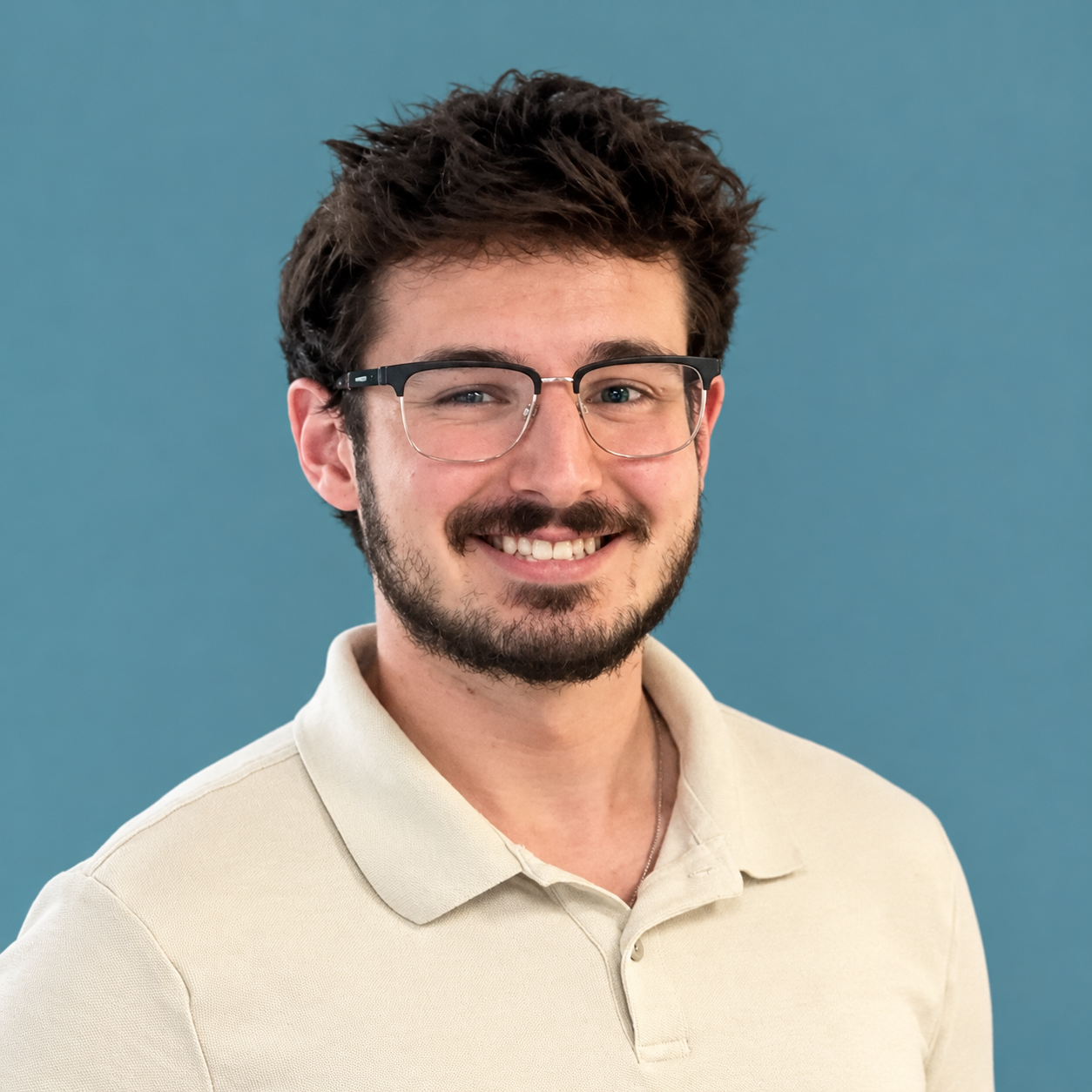A man with glasses, dark hair, and a beard, smiling and wearing a beige polo shirt, standing in front of a blue wall with a colorful abstract painting.