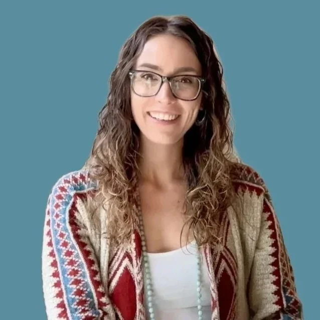 A smiling woman with wavy brown hair and glasses sitting on a white chair in front of a beige wall with astrological and lunar phase artwork.