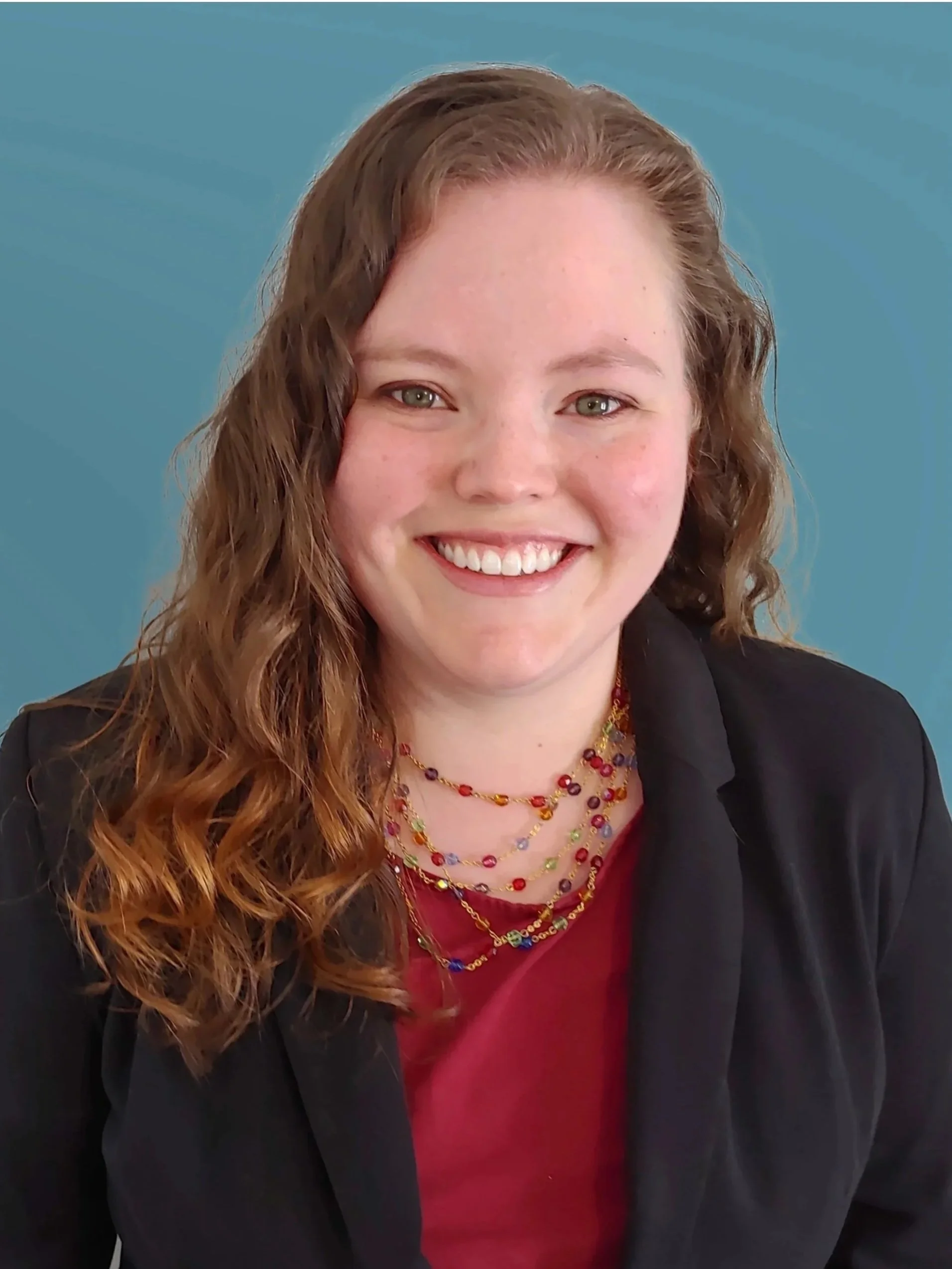 A woman with long, curly red hair smiling, wearing a black blazer over a red top, and several colorful beaded necklaces.
