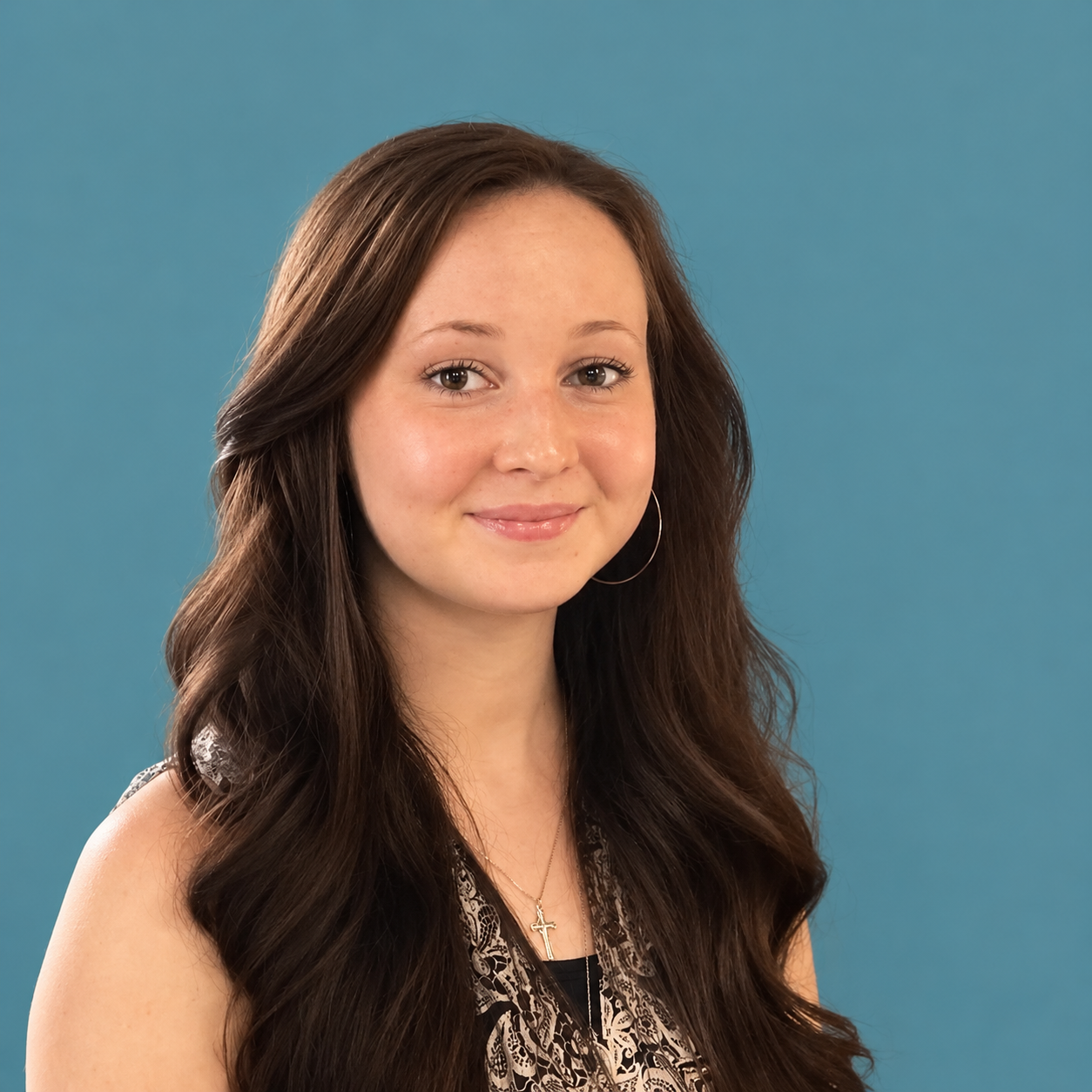 A young woman with long wavy brown hair, wearing a sleeveless top with black-and-white patterns, hoop earrings, and a gold cross necklace, smiling in front of a blue wall with an abstract painting.