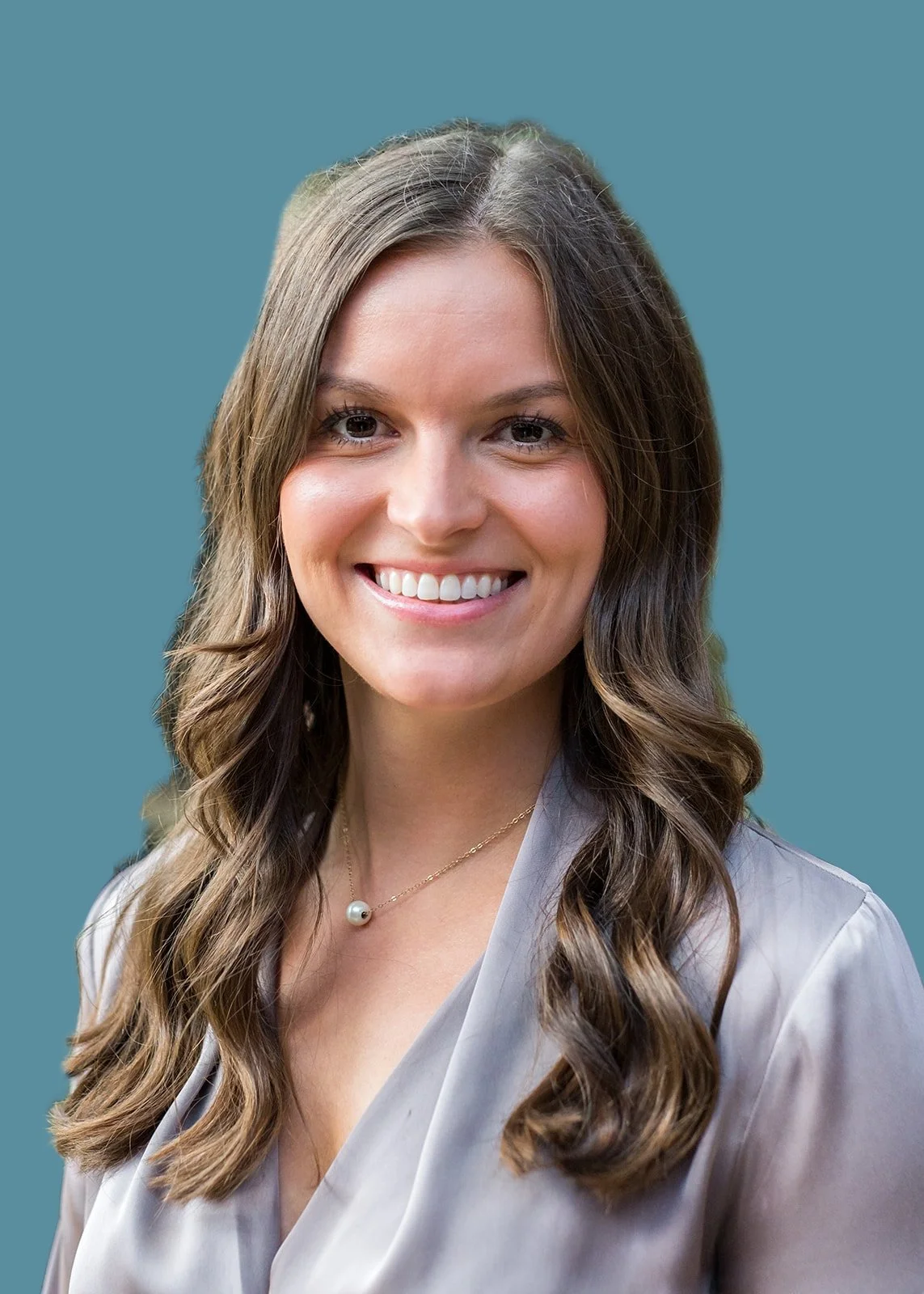 A woman with long wavy brown hair smiling outdoors, wearing a light gray blouse and a delicate necklace with a pearl pendant, with greenery and a brick building in the background.