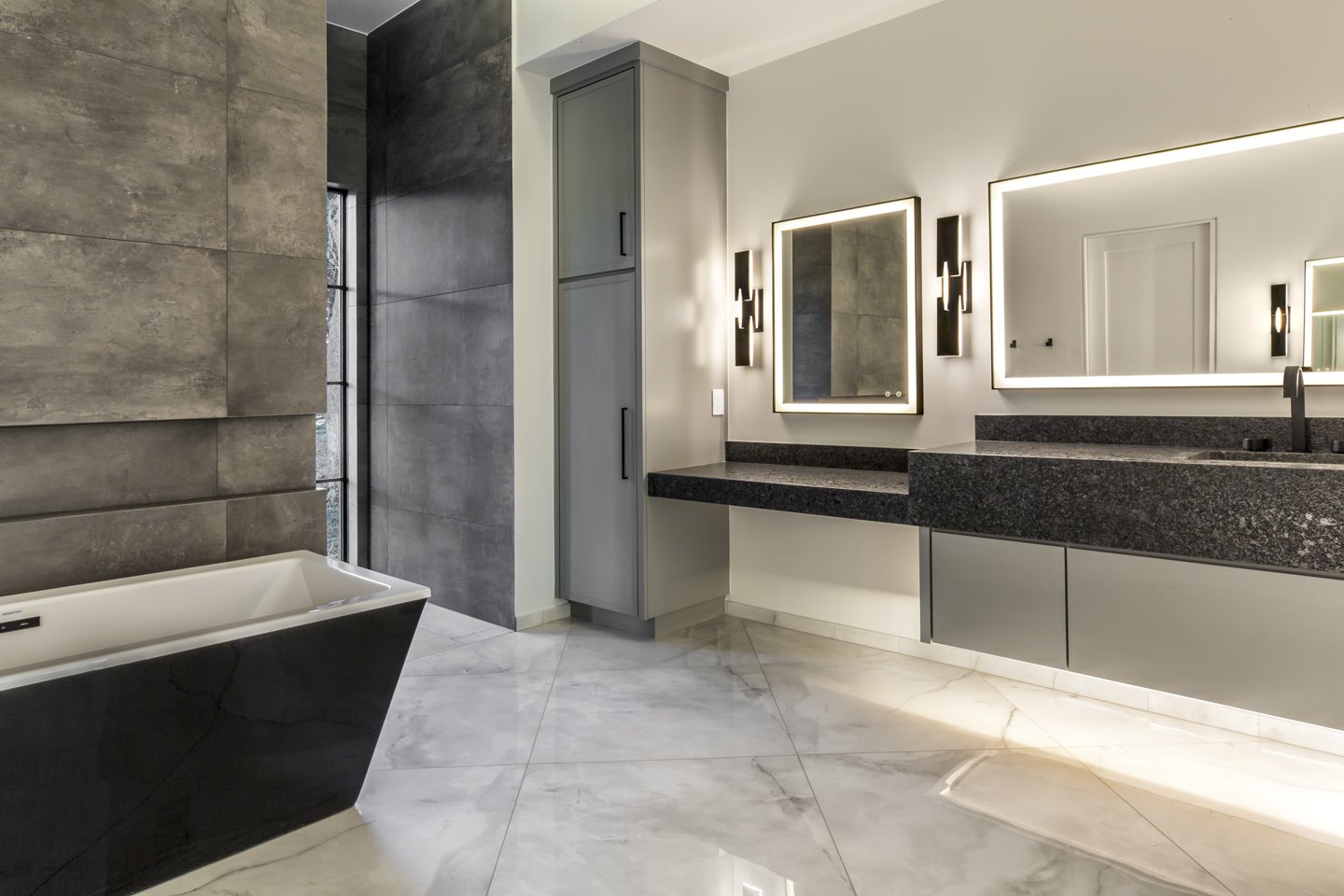 Modern bathroom with gray and black decor, featuring two illuminated mirrors above a black granite countertop, a standalone bathtub, gray cabinets, and marble tile flooring.