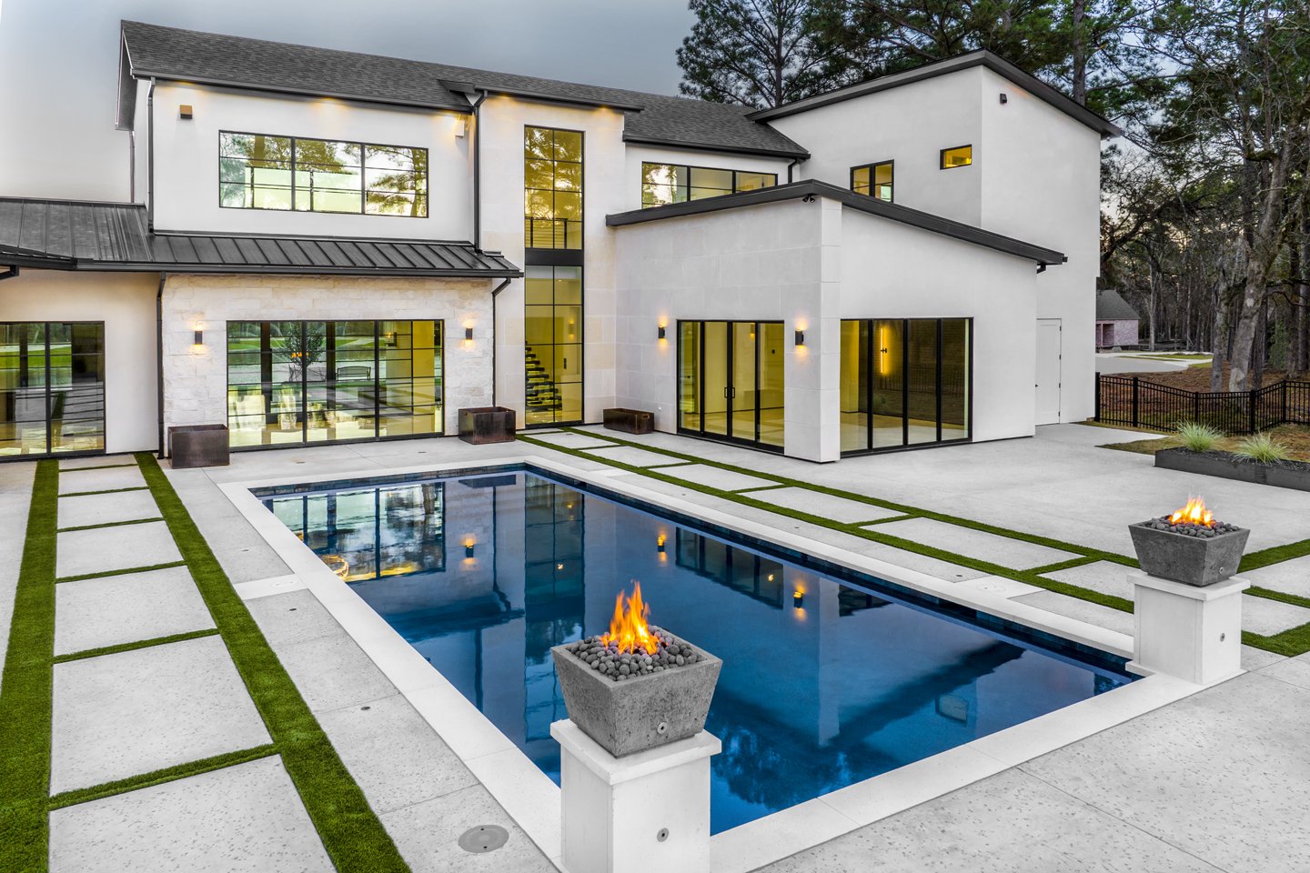 Modern two-story house with large glass windows, a swimming pool with two fire pits, and a spacious concrete backyard, surrounded by trees during dusk.