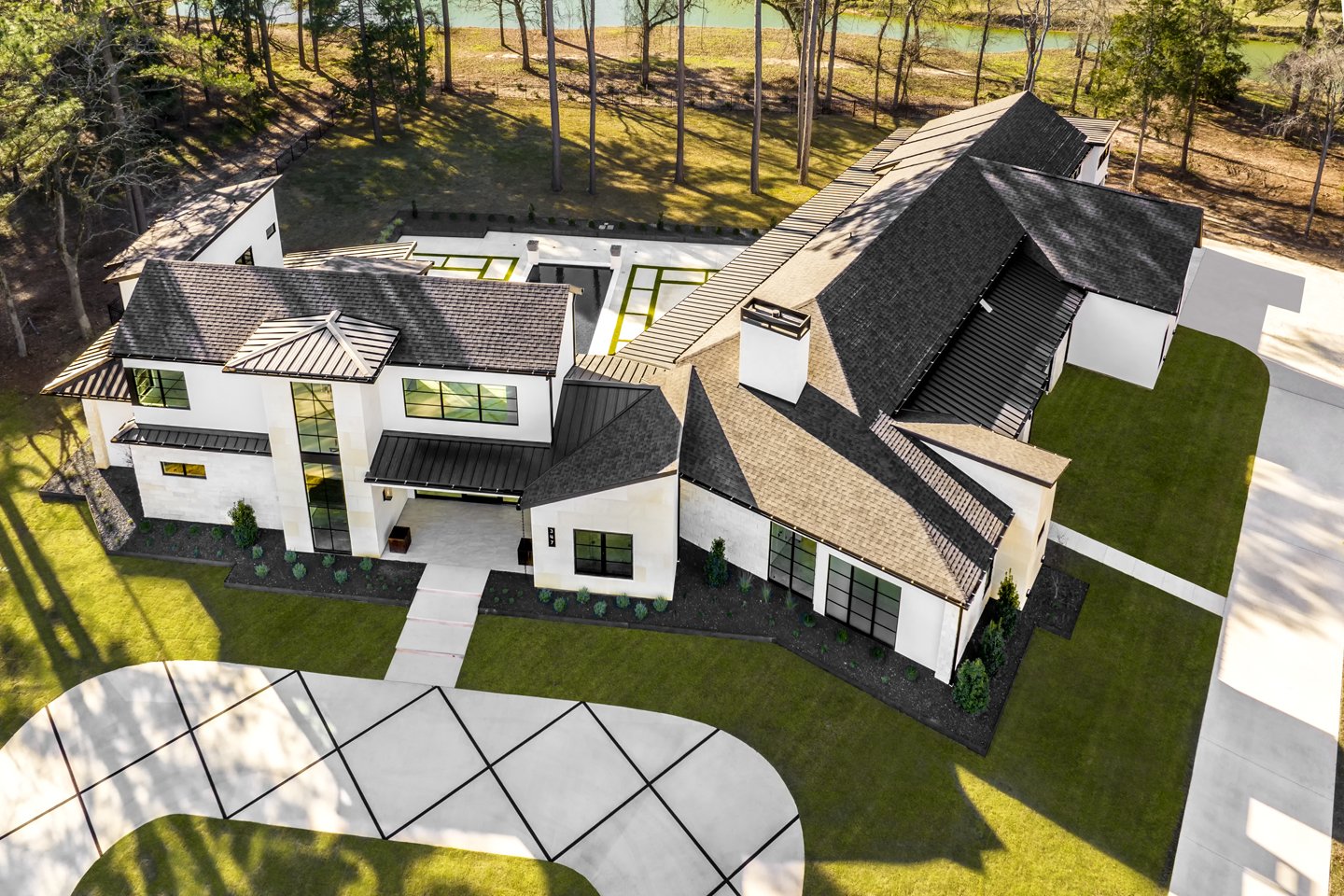 A modern white stucco house with black metal roof, surrounded by green lawn and trees, with a driveway and outdoor patio area.