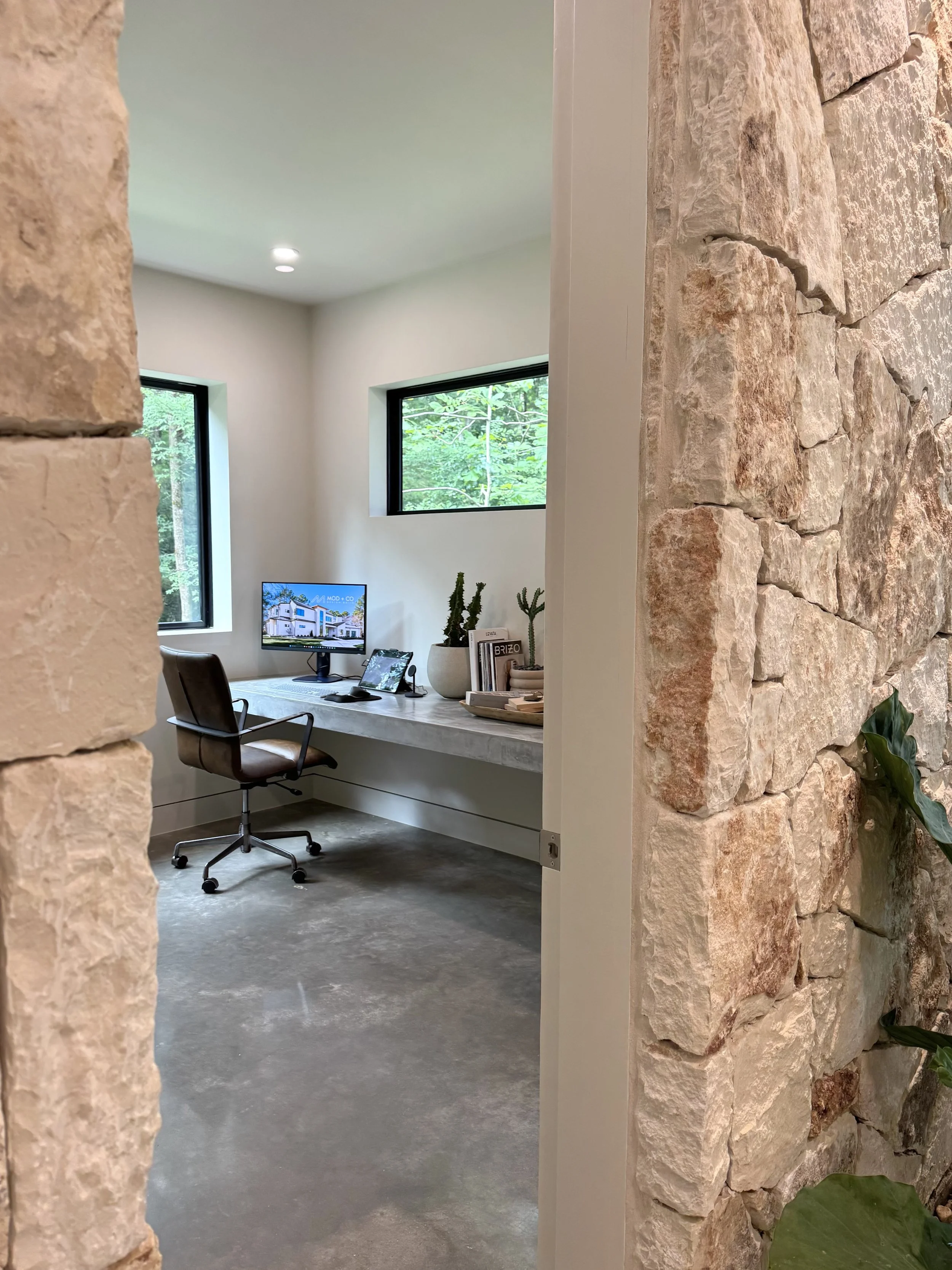 Office with white walls, large windows showing greenery outside, a desk with a computer, books, and decorative plants, and a leather office chair; stone wall partial view in the foreground.