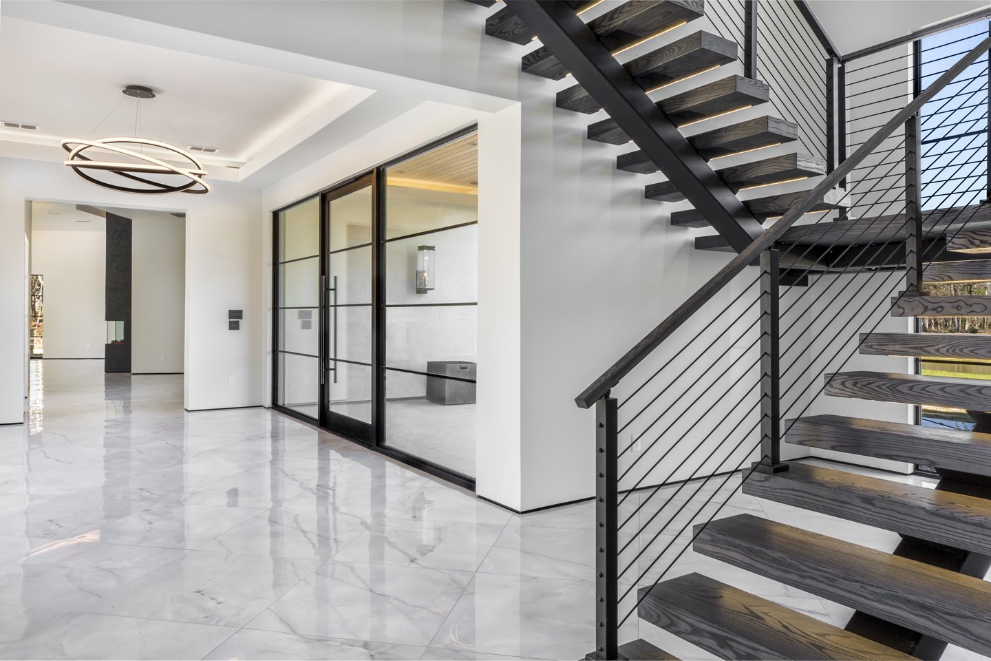 Modern interior with marble floors, black staircase with wood steps and metal railings, glass doors, and minimalist lighting fixtures.