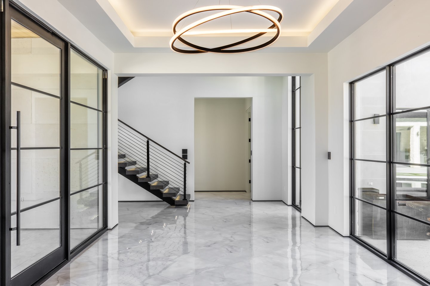 Modern interior space with white walls, marble floors, black-framed glass doors, and a staircase with black railing and wooden steps, illuminated by a circular ceiling light.