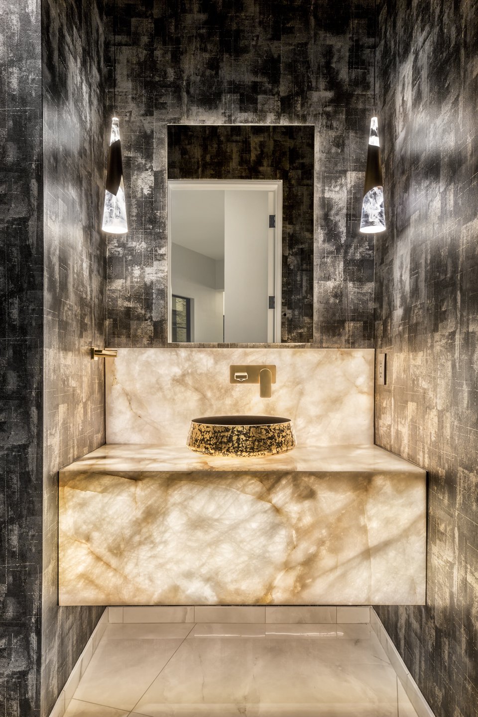 Modern bathroom with black textured walls, backlit onyx vanity with vessel sink, wall-mounted faucet, large mirror, and two black pendant lights.