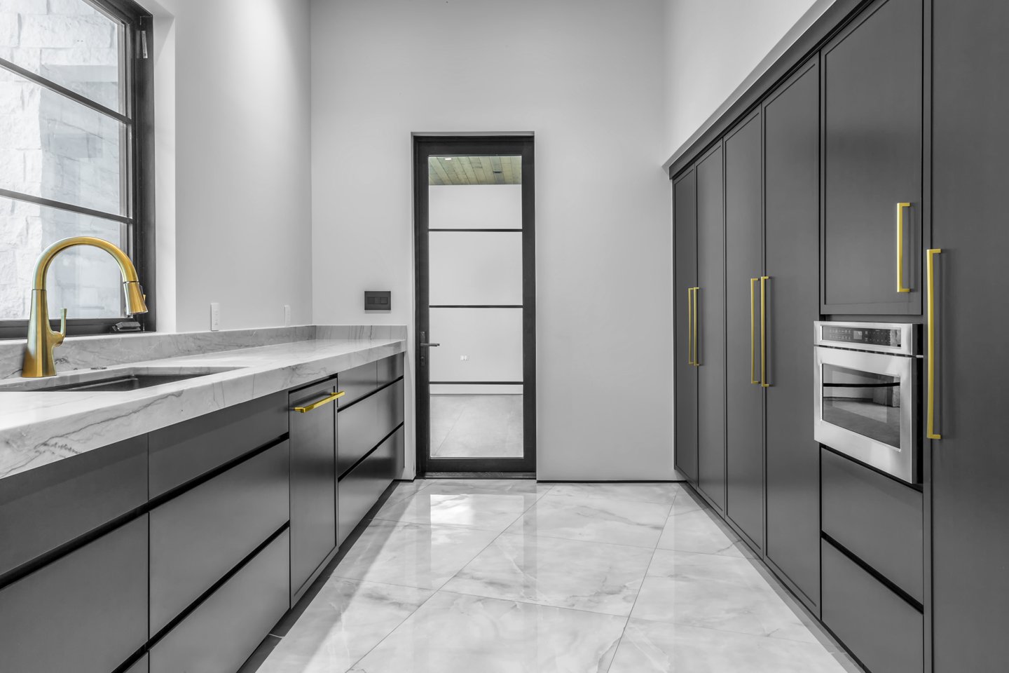 Modern kitchen with dark gray cabinets, gold hardware, marble countertop, gold faucet, marble floor, and a black-framed glass door.