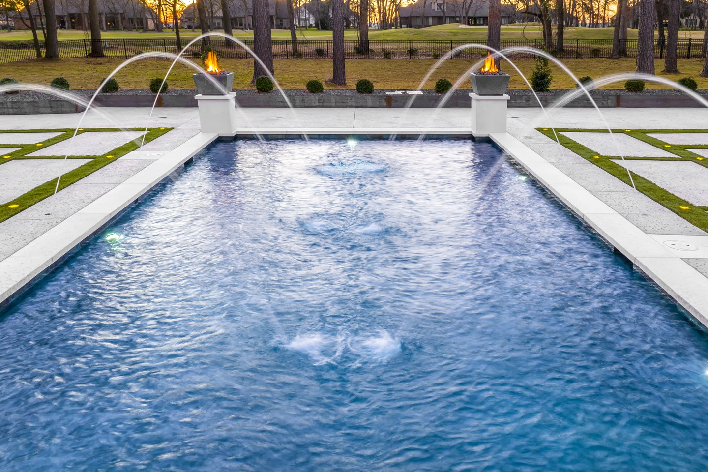 A decorative outdoor pool with water fountains and fire features at the far end, surrounded by a patio area and a fence with trees in the background.