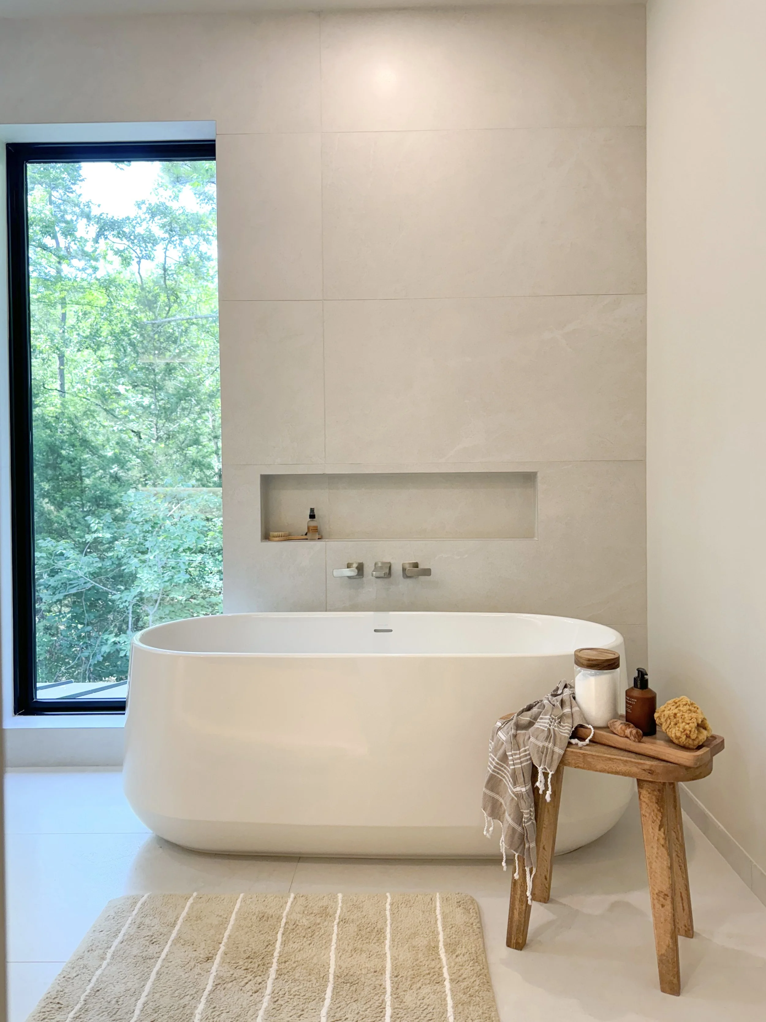 Modern bathroom with freestanding bathtub near large window with green trees outside, beige wall, wooden stool with bath items, and beige bath mat.