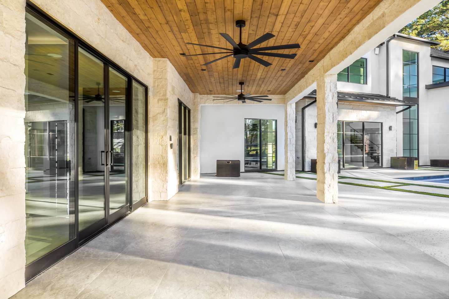 Modern covered outdoor patio area with stone columns, wooden ceiling, ceiling fans, large sliding glass doors, and a view of a swimming pool in the background.