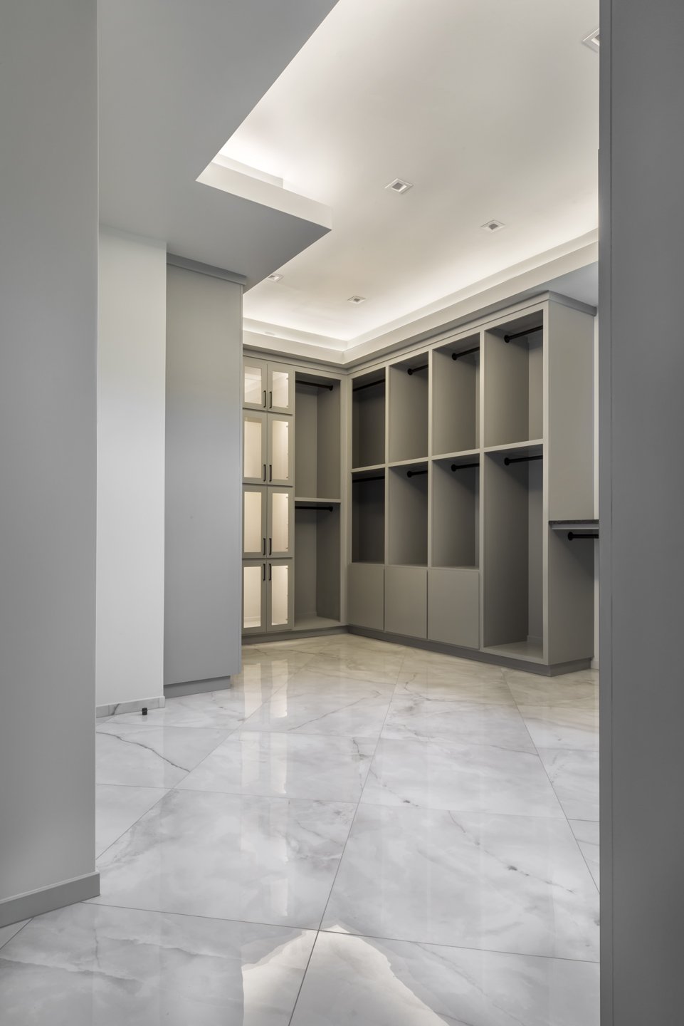 Empty walk-in closet with built-in shelves, drawers, and cabinets, light gray walls, and marble flooring.