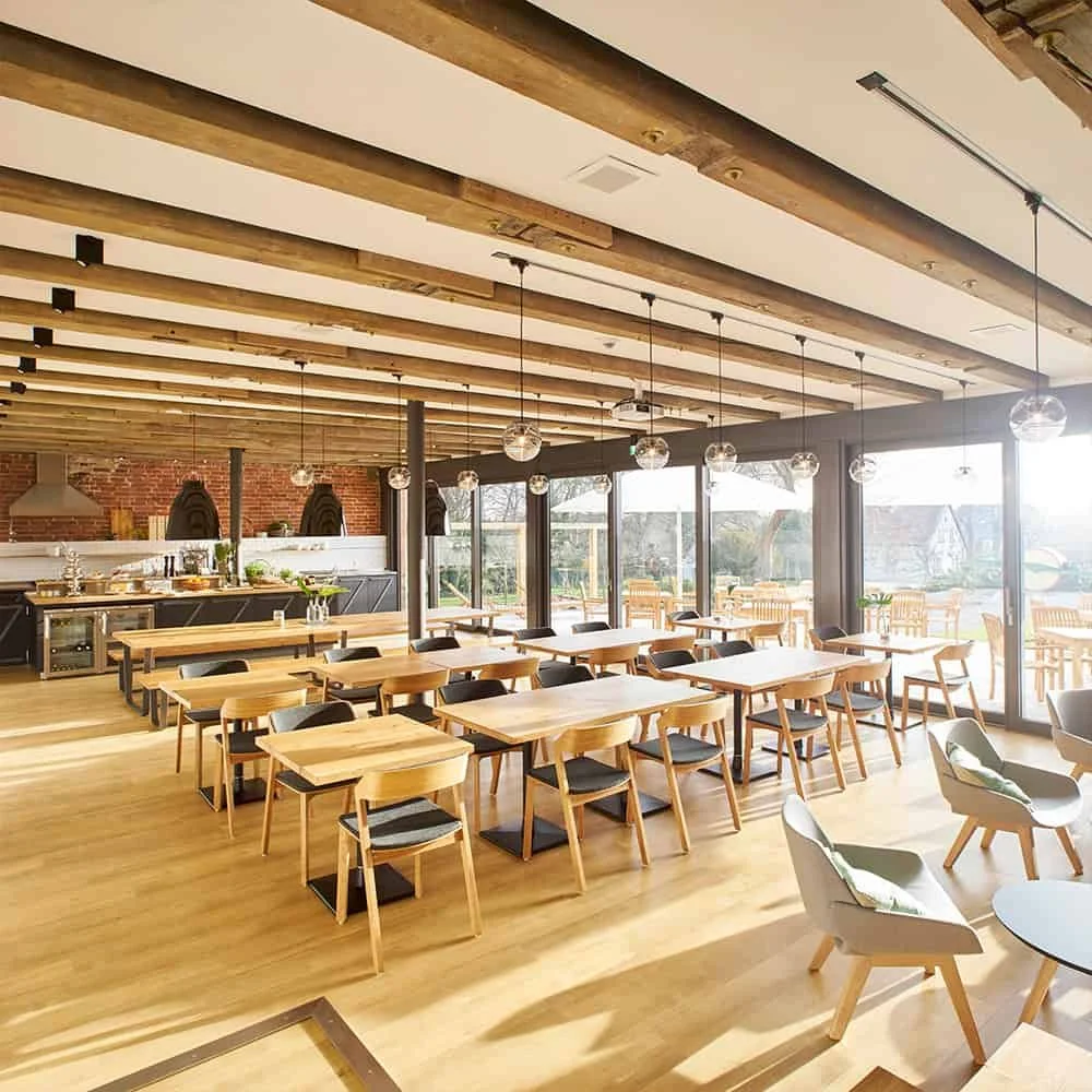 Modern restaurant or cafe interior with wooden tables and chairs, large windows letting in natural light, hanging globe pendant lights, and a visible open kitchen area.