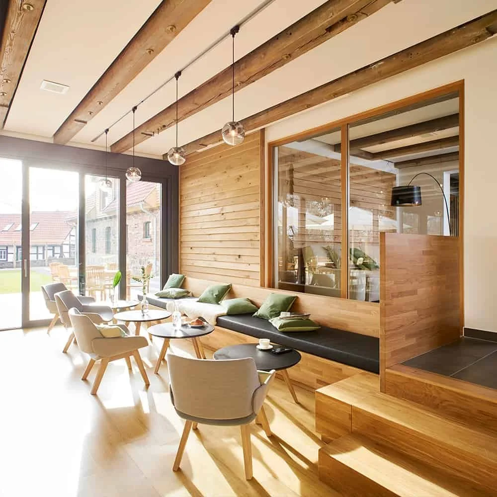 Modern dining area with chairs and tables near large glass doors, wood-paneled wall, and ceiling with exposed wooden beams in a bright space.