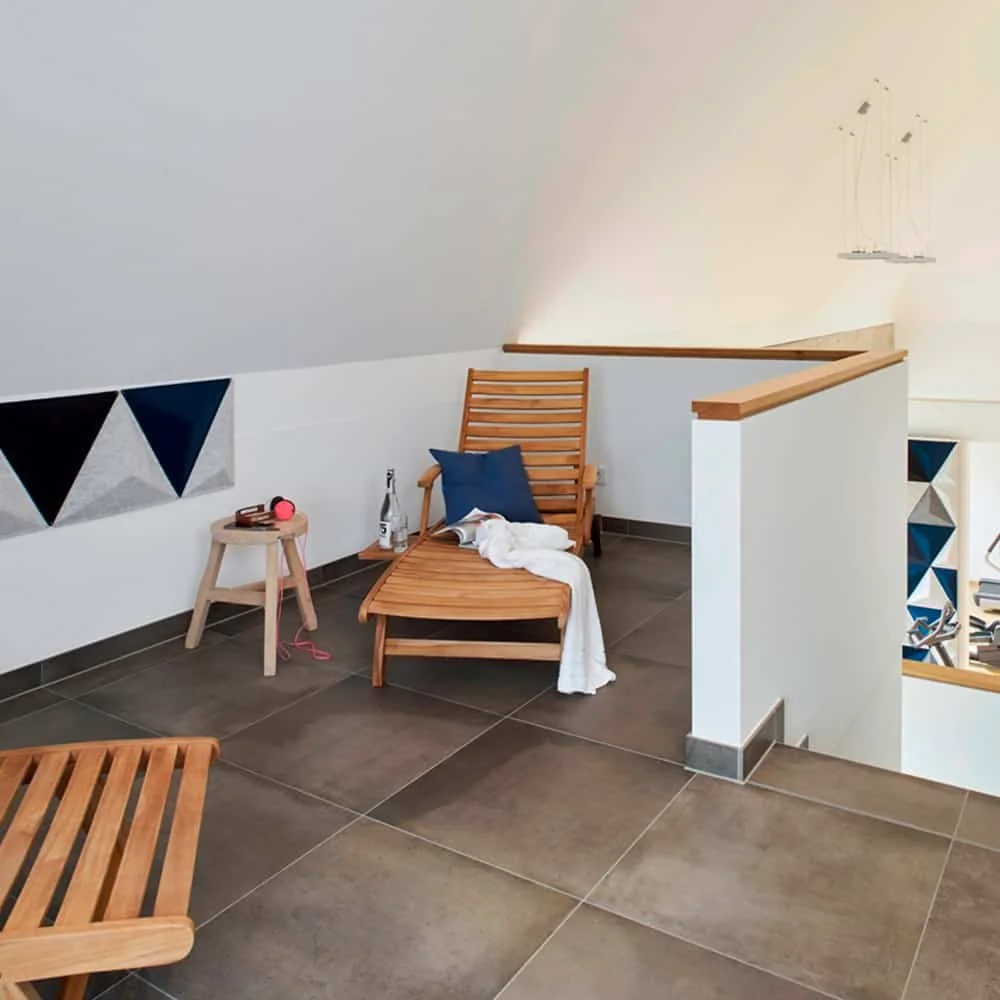 A cozy corner with a wooden lounge chair, a small side table with a bottle and a pink speaker, and decorative wall art in a modern, bright interior.