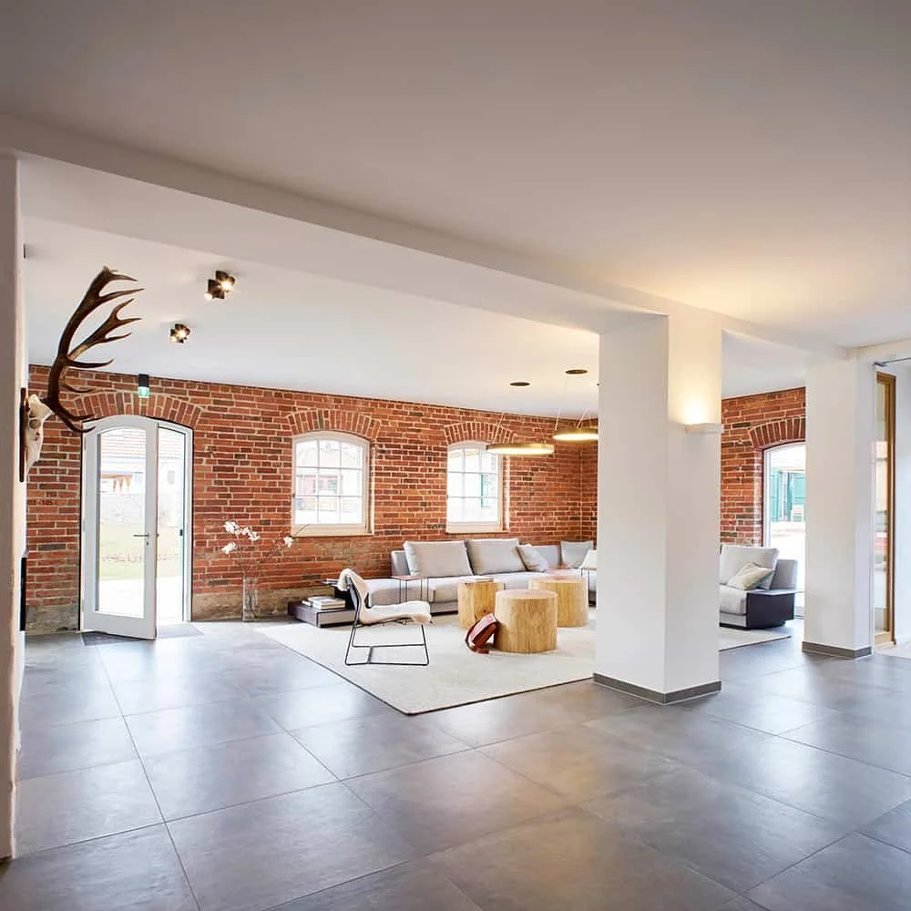 A spacious modern living room with exposed brick walls, large windows, minimalist furniture, and decorative logs on the side table.