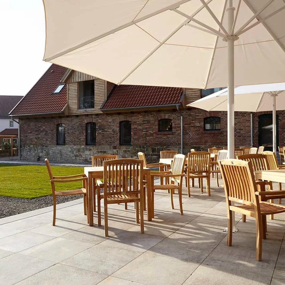 Outdoor patio with wooden chairs and large white umbrellas, in front of a brick building with a red tiled roof.