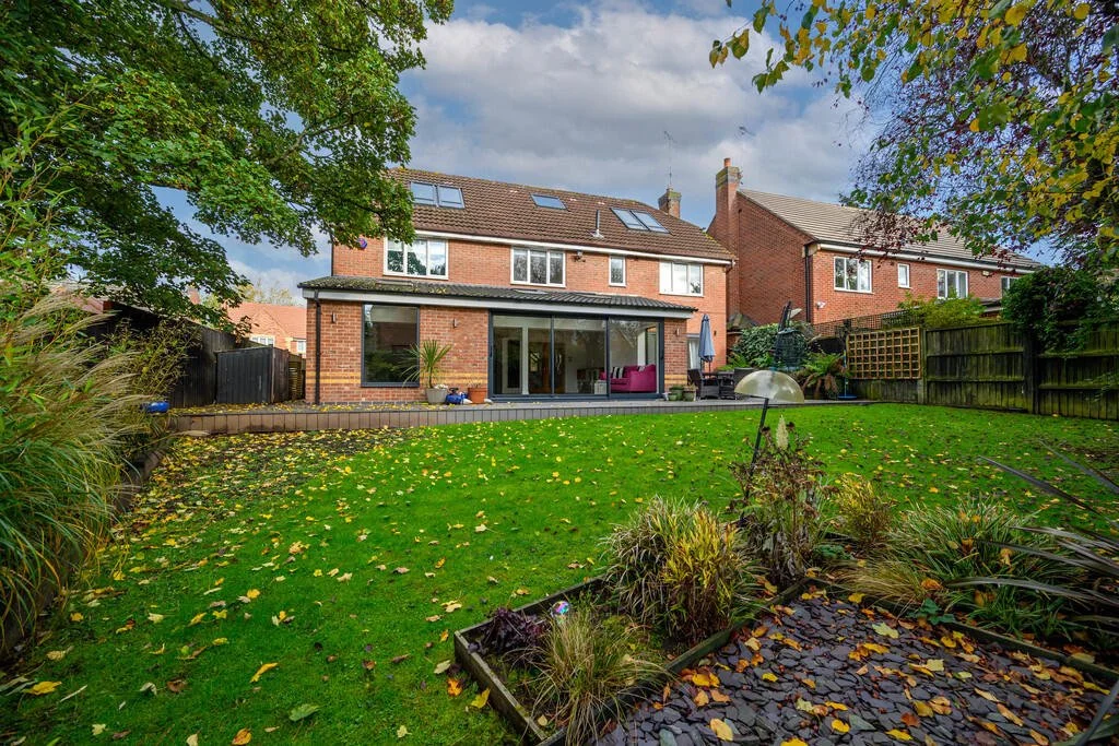 Merritt Estates. Backyard of a brick house with large glass sliding doors, patio furniture, and a lush green lawn with fallen yellow leaves.