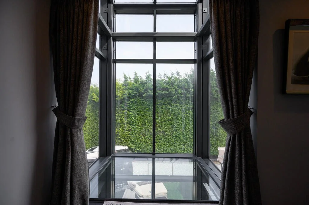 Merritt Estates. A window with a glass floor view, framed by dark curtains, overlooking green trees outside.