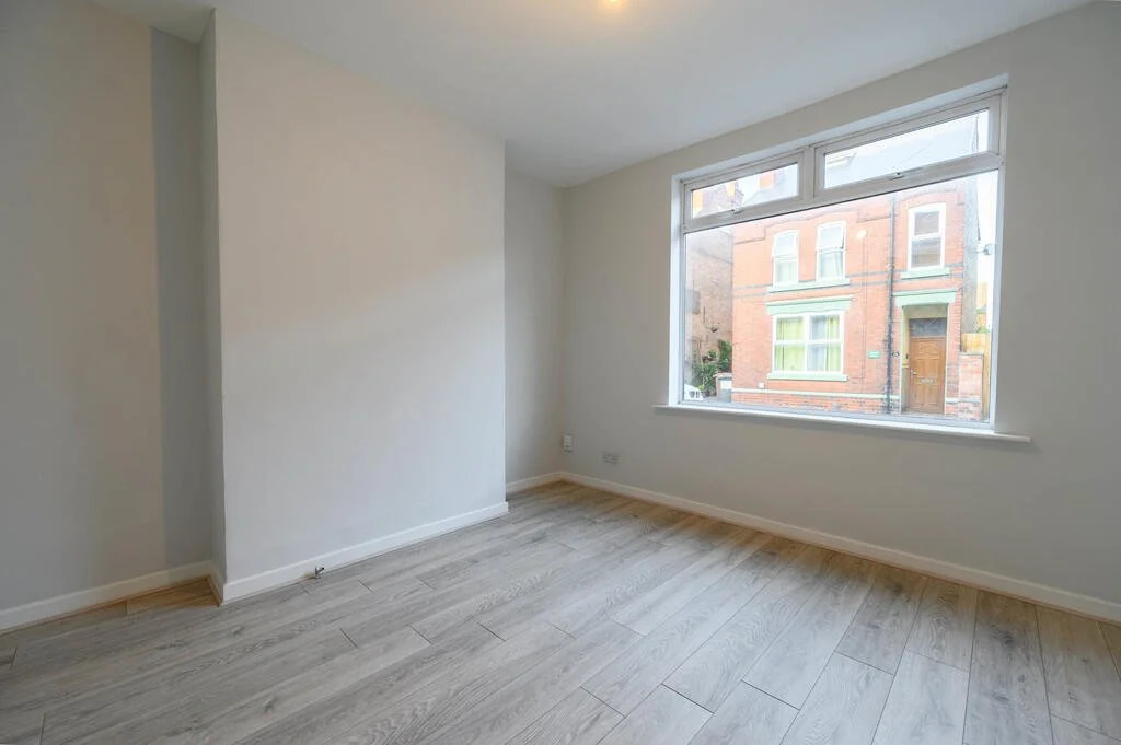 An empty room with white walls, light wood flooring, and a large window showing a view of neighboring brick buildings.