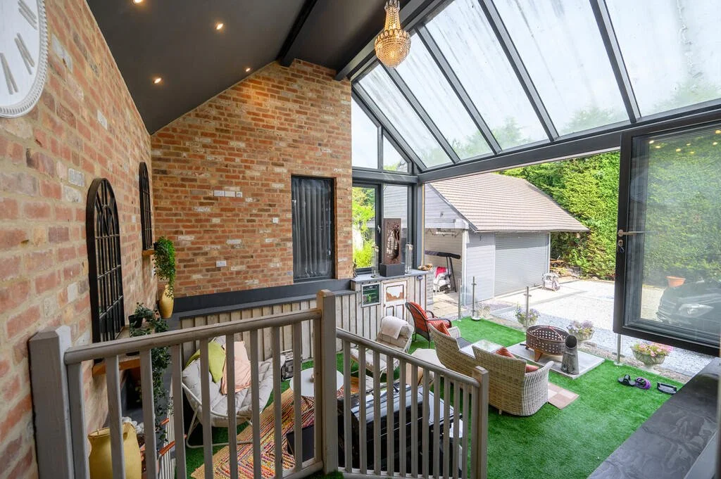 Merritt Estates. Sunroom with brick walls, large glass windows and ceiling, patio furniture including wicker chairs and a small table, green artificial grass flooring, and a view of a backyard with a garage and trees.