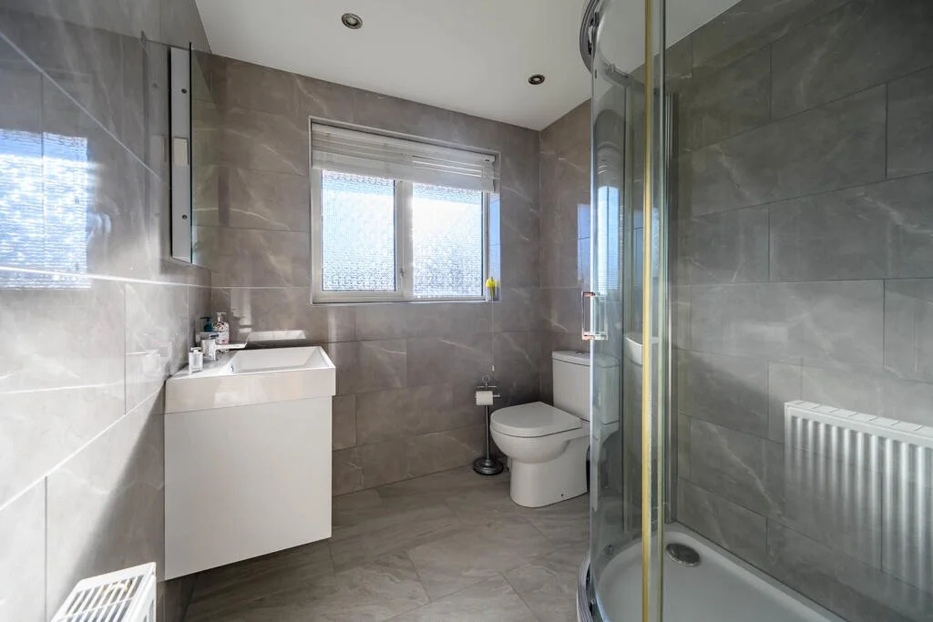 A modern bathroom with grey tiled walls and floor, a white vanity with toiletries, a toilet, a window with blinds, and a glass-enclosed shower.