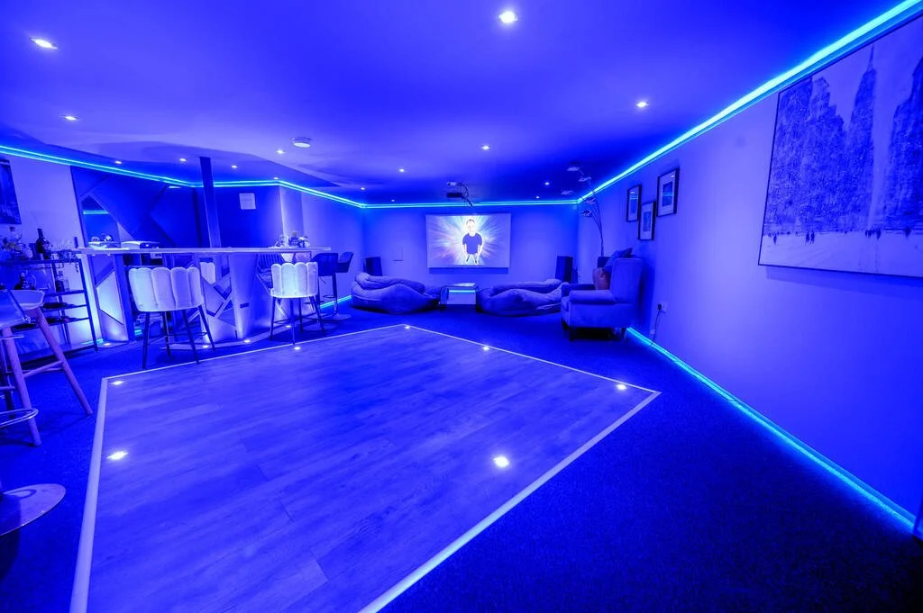 Merritt Estate. A dimly lit basement or entertainment room with blue LED lighting along the ceiling and walls, a small dance floor outlined with LED lights, a bar area with barstools, bean bag chairs, a large screen TV, and framed artwork on walls.