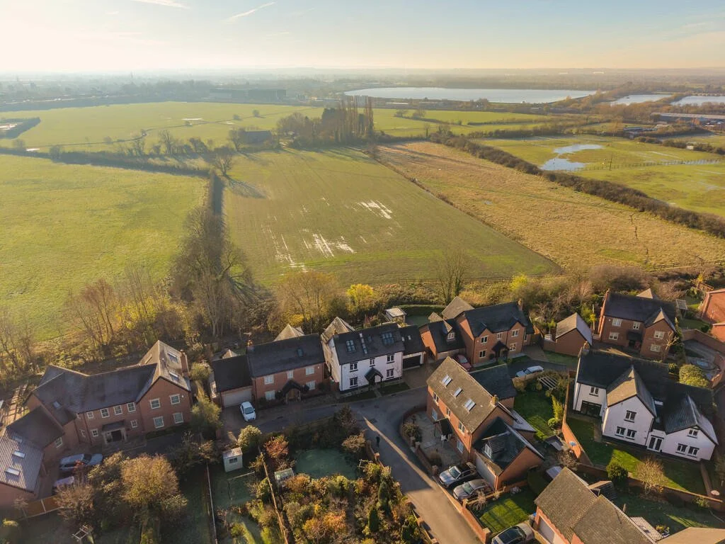 Aerial view of a suburban neighborhood with houses and gardens in the foreground, and expansive green fields and ponds in the background, under a clear sky.
