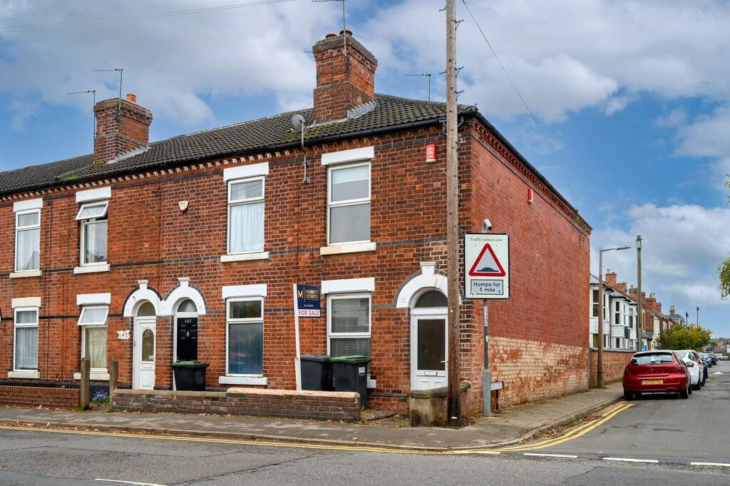 Merritt Estates. A row of terraced brick houses on a residential street with cars parked along the curb. There are street signs, including one warning about humps for 1 mile, and a 'for sale' sign on one house.