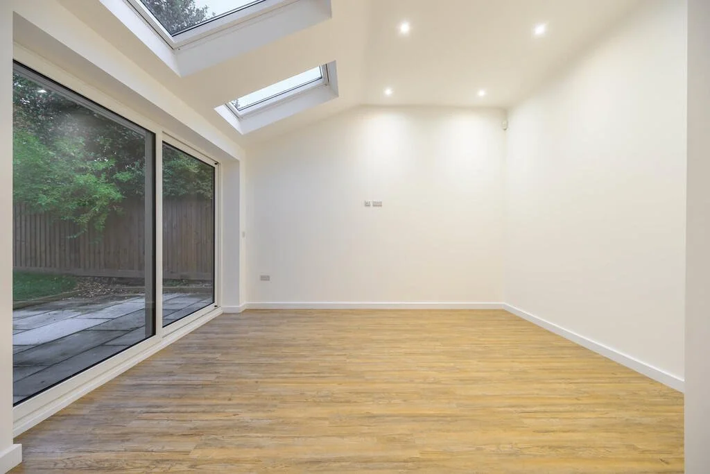 Merritt Estates. An empty room with wooden flooring, white walls, and large sliding glass doors leading to a backyard, with skylights on the ceiling bringing in natural light.