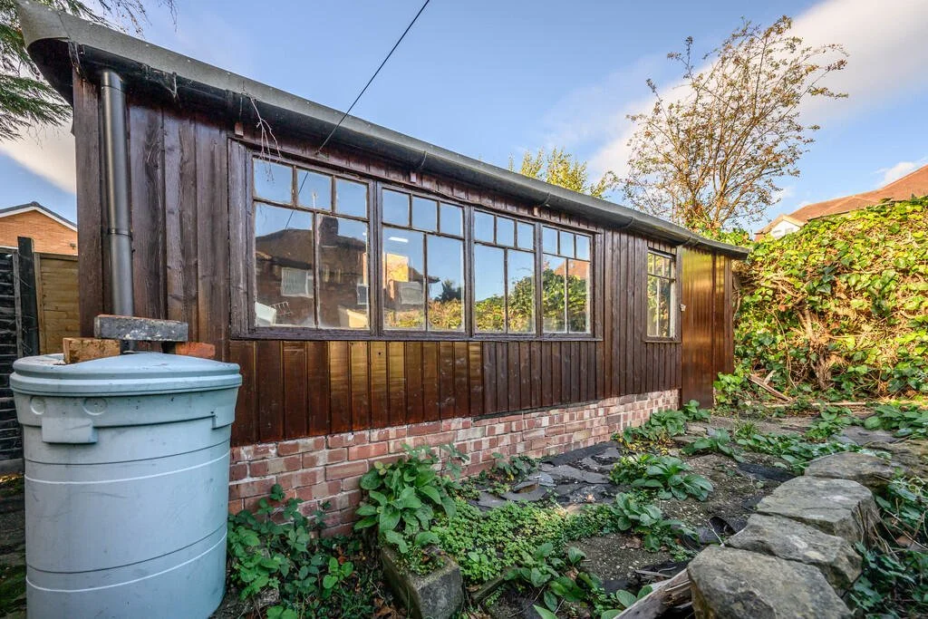Merritt Estates. Backyard garden with a small wooden shed, brick foundation, overgrown plants, and a large grey rain barrel on the left.