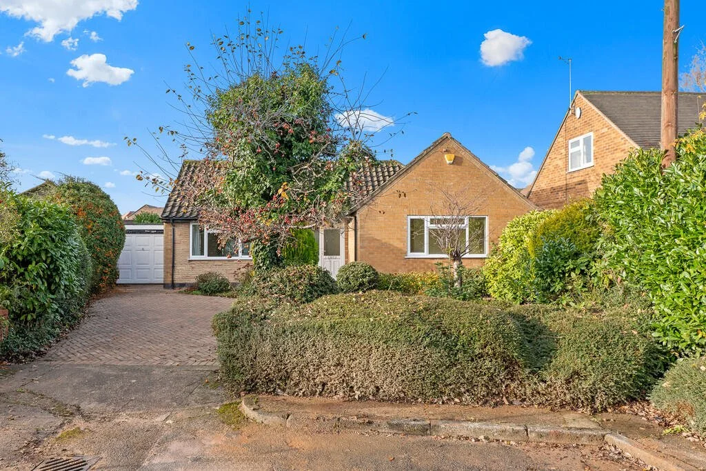 Front view of a single-story brick house with a garden, driveway, and garage, under a bright blue sky with some clouds.
