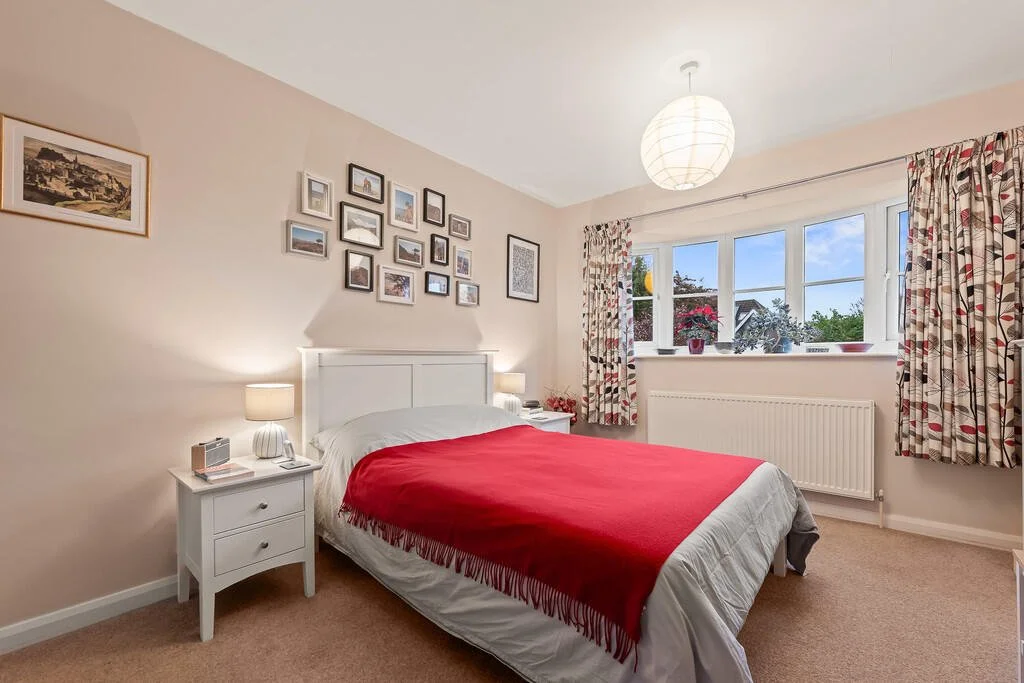 A bedroom with a white bed, red blanket, nightstands with lamps, and a window with curtains showing a house and trees outside.