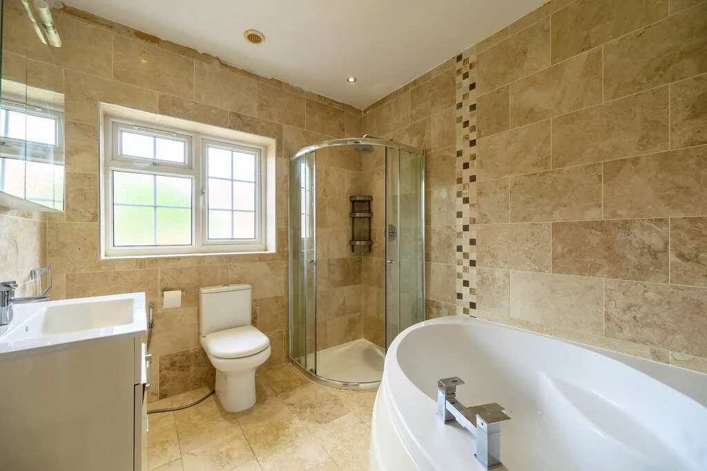 Modern bathroom with beige tile walls and floor, a white sink vanity, a toilet, a corner shower with glass doors, a large frosted window, and a corner bathtub.