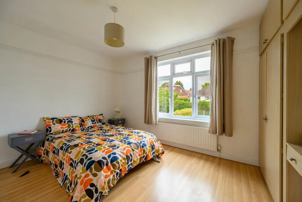 A bedroom with a bed covered in a multicolored geometric patterned duvet, a nightstand with a lamp, a large window with beige curtains, a ceiling light fixture, and built-in wardrobe cabinets.
