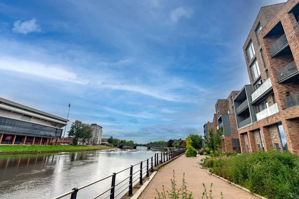 View of a river with a walkway on the right side, modern apartment buildings with balconies, and a bridge in the distance under a partly cloudy sky.