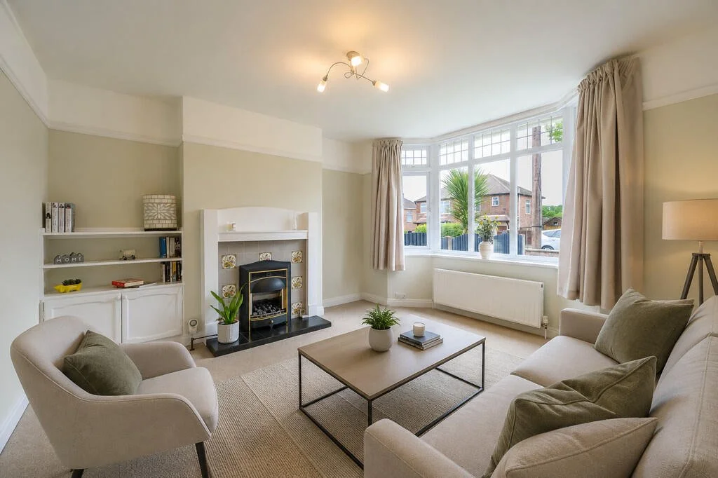 Bright living room with large bay window, beige sofa, armchair, fireplace, built-in shelves, potted plants, and neutral decor.