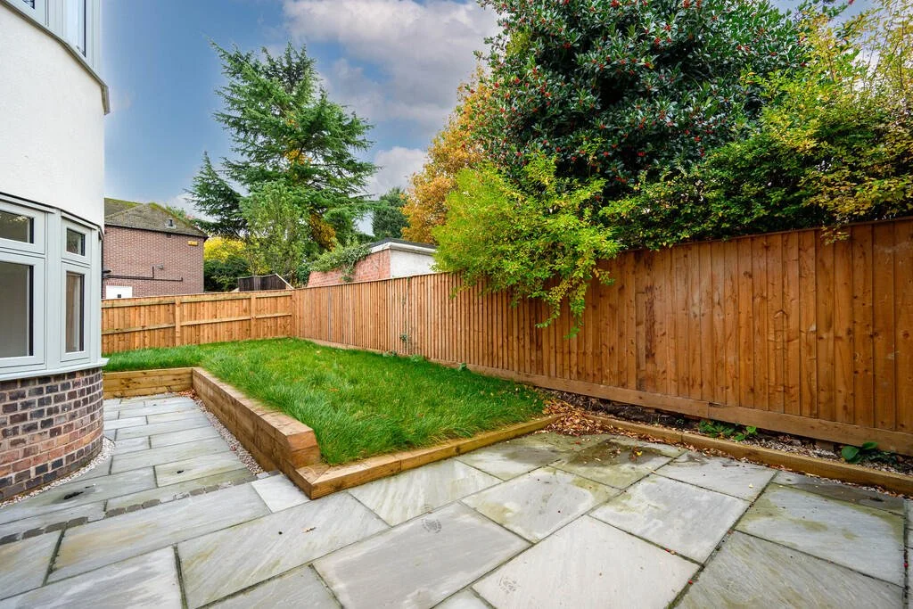 Merritt Estates. A backyard with a small grassy lawn, a fenced perimeter, and a paved patio area next to a house with brick and white siding. There are trees with green and some yellow leaves, indicating the fall season.