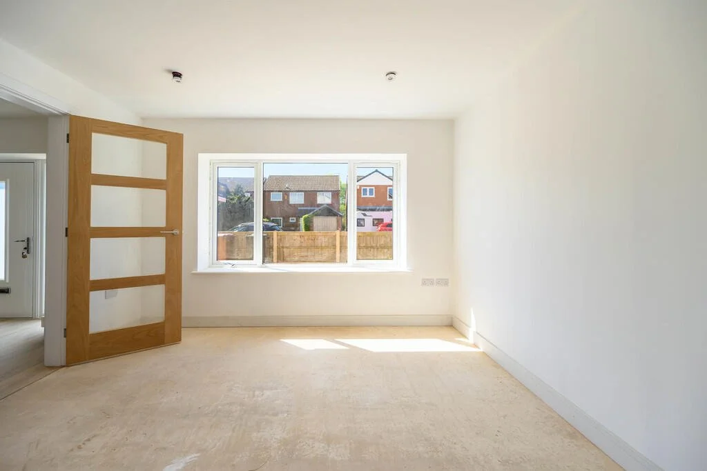Empty room with white walls, carpeted floor, a wooden door, and a large window showing houses outside.