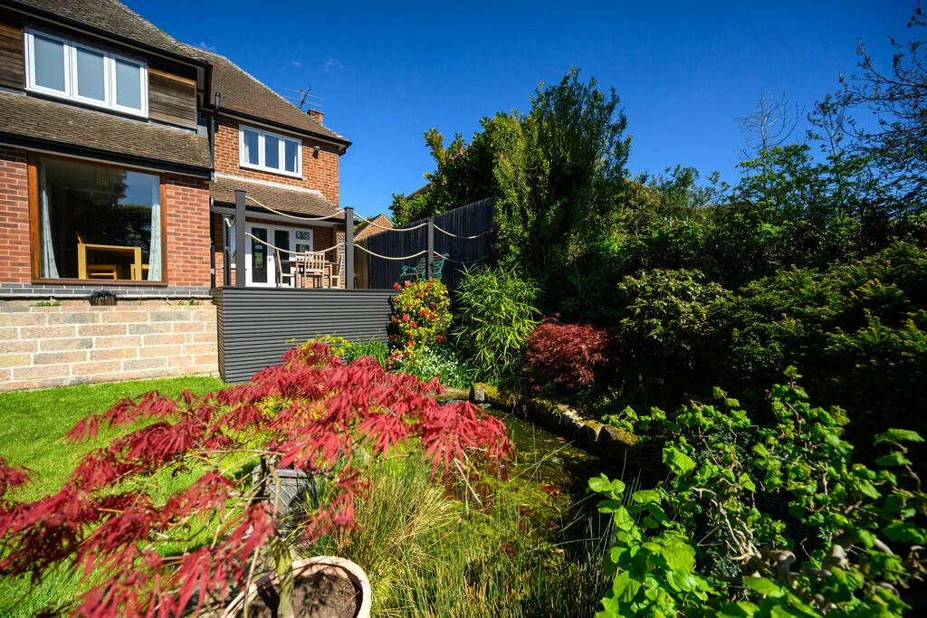 A backyard garden with a raised deck, patio furniture, lush green plants, and red and green foliage under a clear blue sky.