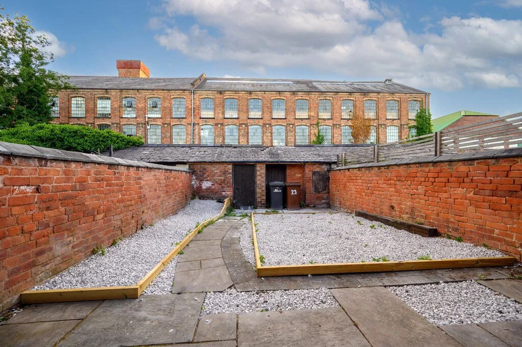 Merritt Estates. Backyard with gravel, brick walls, and a paved pathway leading to a door, with a large brick building in the background under a partly cloudy sky.