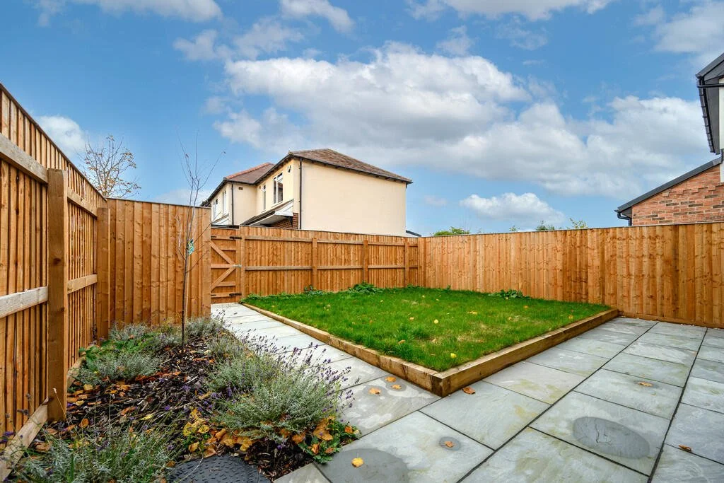 Merritt Estates. Backyard with a wooden fence, small tree, flowering plants, a grassy patch, and a paved patio area under a partly cloudy sky.