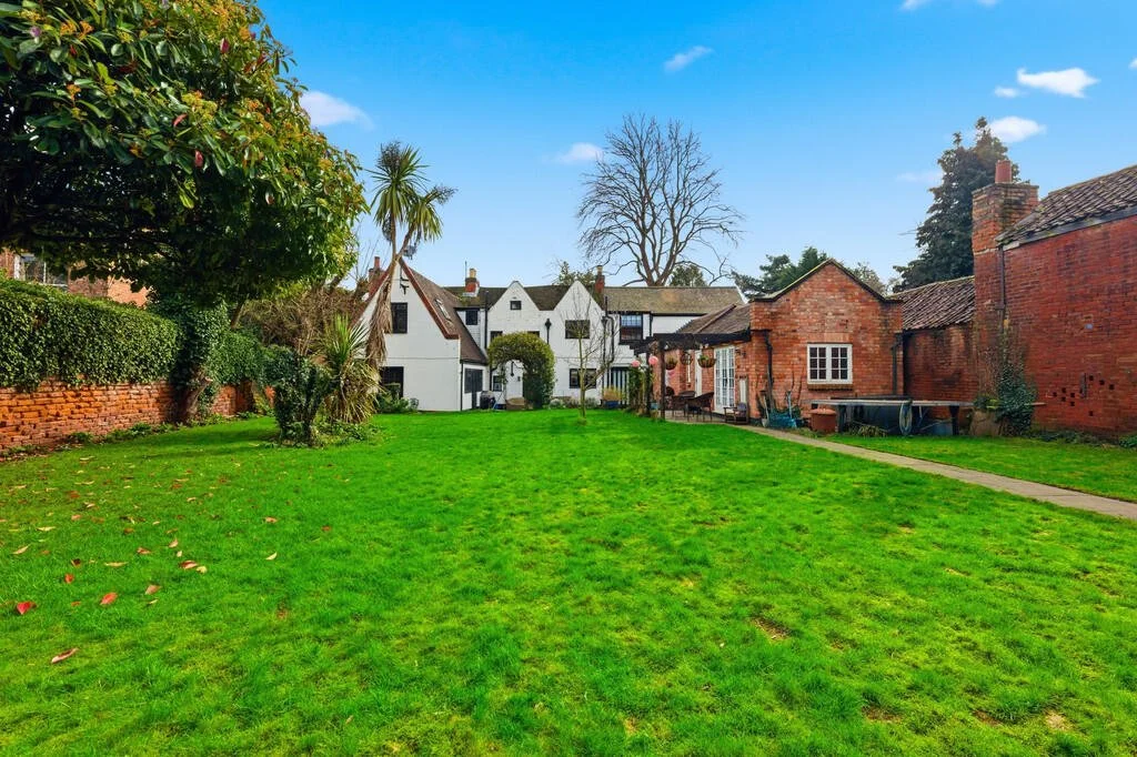 A lush green backyard with a large grass lawn, trees, and shrubs. In the background, there are charming white and brick houses with a bright blue sky above.