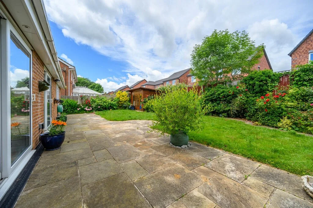 Backyard patio with stone tiles, green grass, and blooming bushes, with a blue sky and clouds.